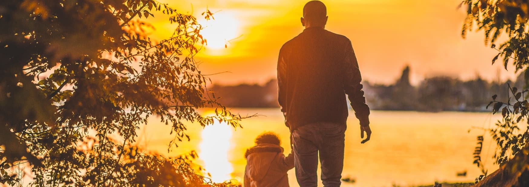 A parent and young child walking hand in hand toward a sunset over the water