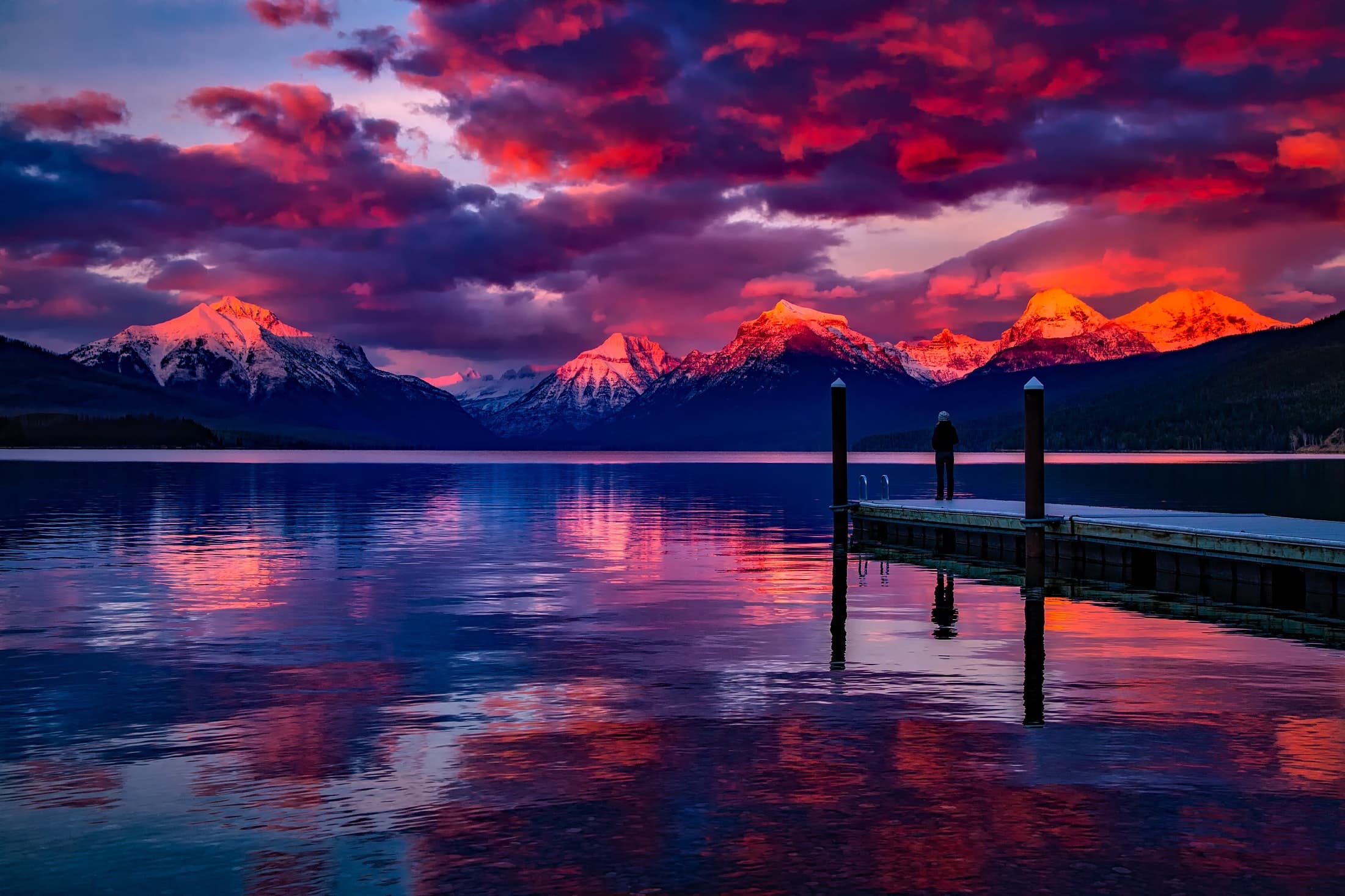 Snow-capped mountains glowing at dusk above a still lake, a lone figure standing on a dock